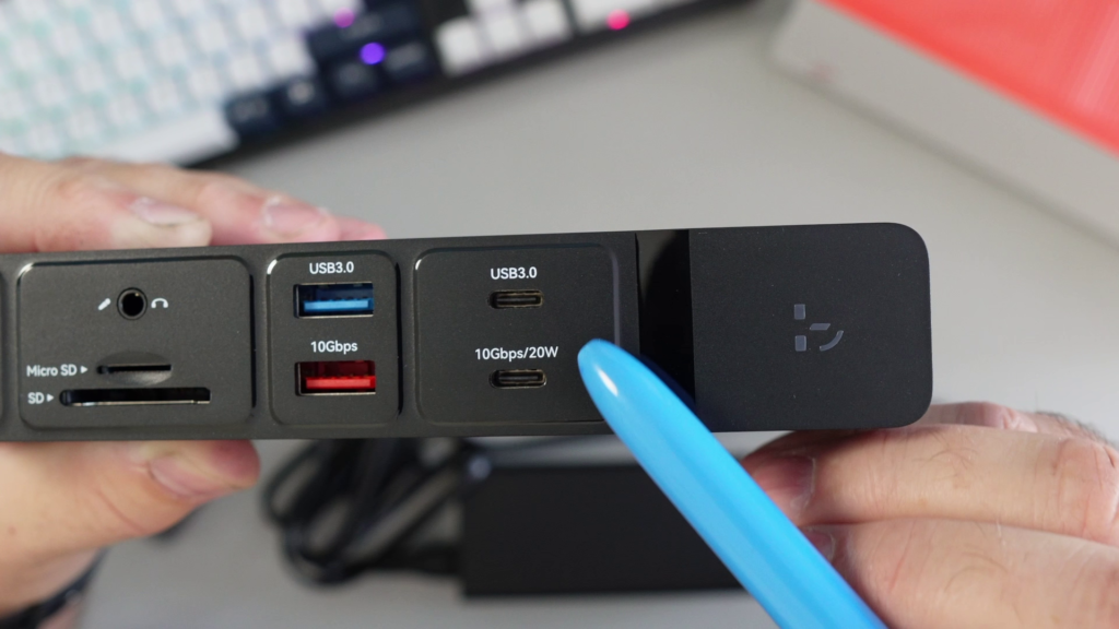A close-up of the front panel of the TobenONE UDS045 docking station, showing SD/micro SD slots, audio jack, two 10Gbps USB-A ports (one blue, one red), one 10Gbps/20W USB-C port, and the power button, with a blue pen pointing to the USB-C port.