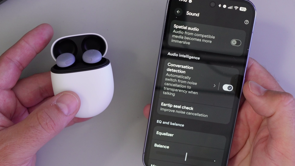 A photo showing the Google Pixel Buds Pro 2 case next to a Pixel phone displaying the "Conversation detection" setting enabled in the Sound menu.