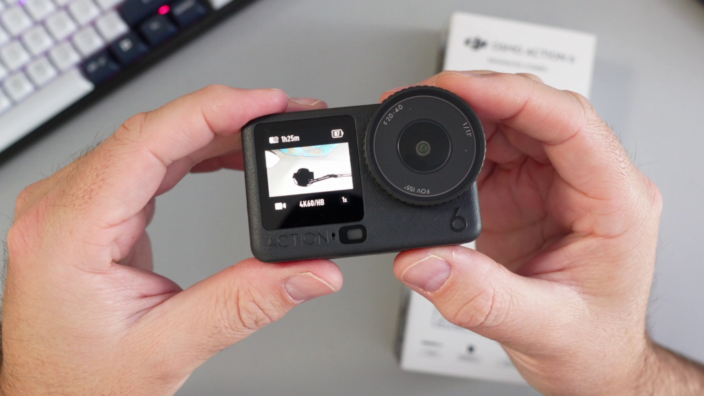 A close-up of hands holding the DJI Osmo Action 6 camera, showing the active front touchscreen display and the large lens with f/2.0-4.0 variable aperture markings.