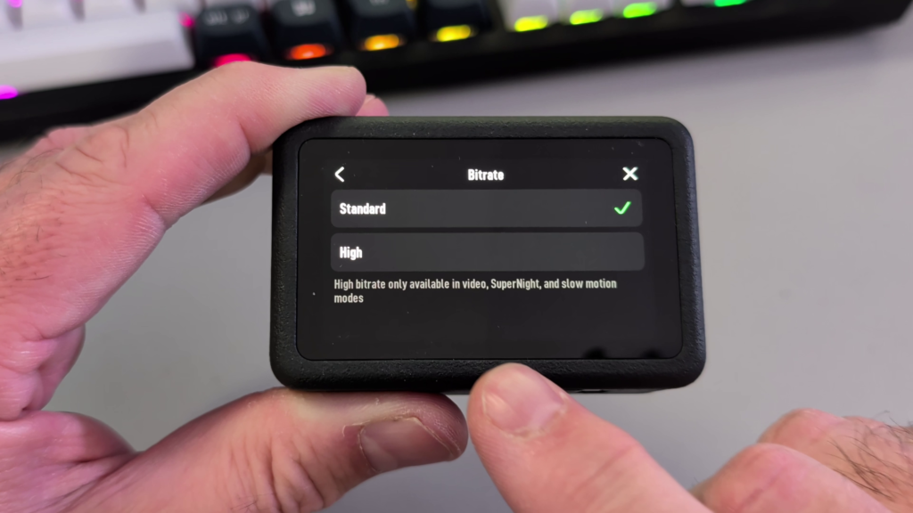 Hands holding a DJI Osmo Action 6 camera, displaying the Bitrate menu screen where the green checkmark is currently next to the default 'Standard' option.