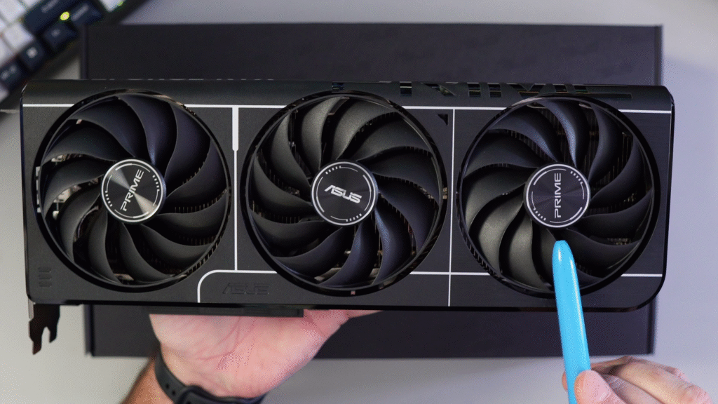 A close-up view of the ASUS Prime GeForce RTX 5060 Ti graphics card held in hand, showing the triple-fan cooling design. A blue tool points to the rightmost fan hub featuring the PRIME logo.