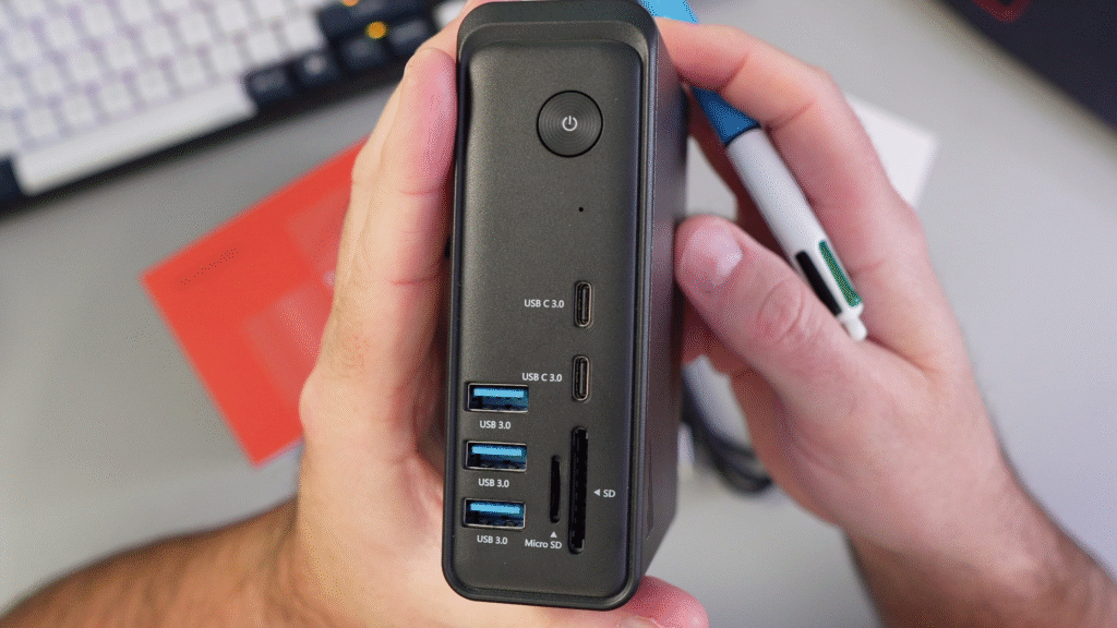 A close-up photograph of the black TobenONE UDS057 docking station being held upright by two hands. The front panel is clearly visible, showing a circular power button at the top, followed by two USB-C 3.0 ports, three USB-A 3.0 ports, a Micro SD card slot, and a full-size SD card slot at the bottom. A hand holding a multi-color pen is on the right.