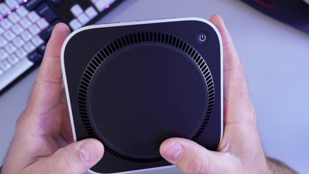 A close-up view of the bottom surface of the black M4 Mac Mini, highlighting the relocated power button near the rear corner. A cooling vent is also partially visible on the underside.